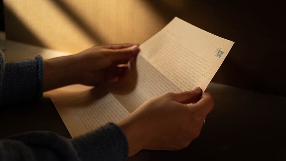 Unas manos sostienen abierta una carta de Otro Aire, iluminada por la luz de una ventana