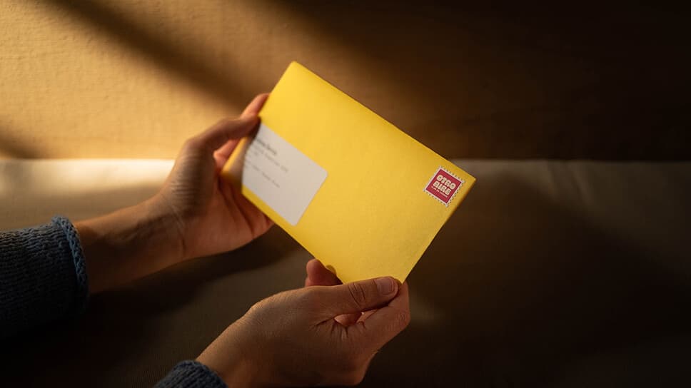 Unas manos sostienen el sobre amarillo de Otro Aire, cerrado, con la etiqueta del destinatario y la estampilla coral del club